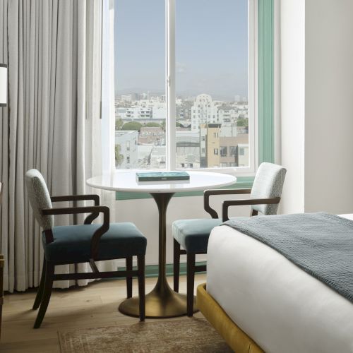 A hotel room with a bed, round table, two chairs, and city view through a window. A bar area with drinks on a countertop is visible.
