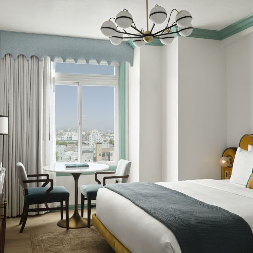 A modern hotel room with a double bed, stylish decor, small table with chairs, and a large window with city views.