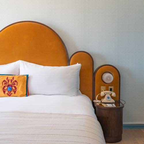 A modern bedroom features a mustard headboard, white bedding, decorative pillow, and a vintage phone on a wooden nightstand.