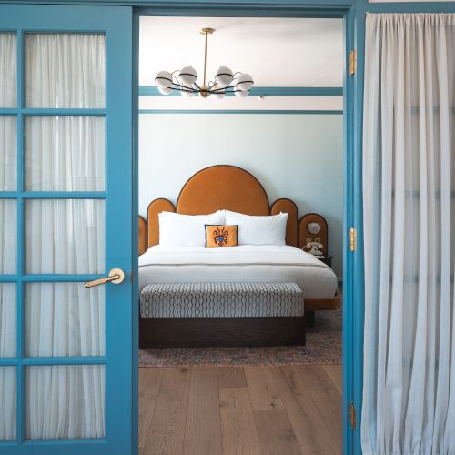 A room with blue-framed glass doors opens to a cozy bedroom with a double bed and wooden floor, lit by an overhead chandelier.