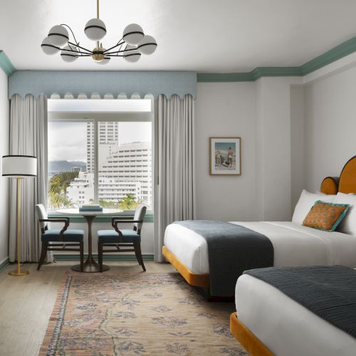 The image shows a hotel room with two beds, a large window with a view, a table with chairs, and stylish lighting and decor.