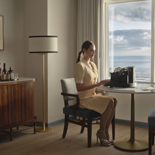 A person sits at a table with a typewriter in a room overlooking the ocean. There's a cabinet with drinks and a lamp nearby.