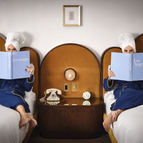 Two people in robes and towels on heads reading "Los Angeles" books, sitting on beds next to each other with a vintage phone.