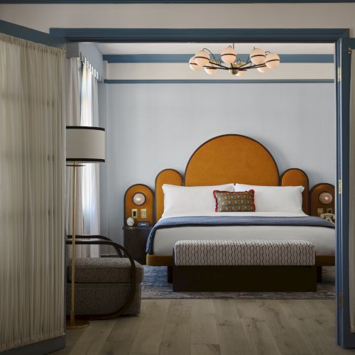 A stylish bedroom with an orange headboard, double doors, floor lamp, and chandelier, featuring a cozy bed and modern decor.