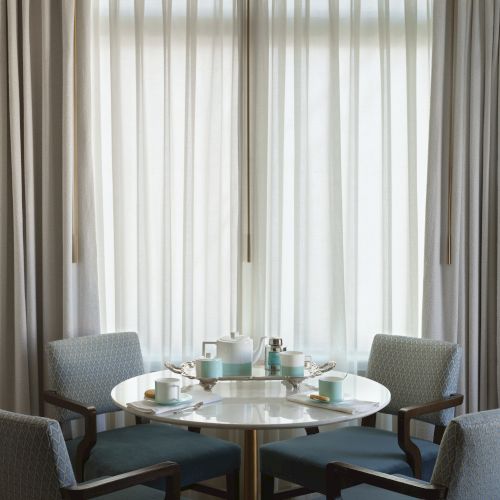 A round table with a tea set and four chairs is in front of sheer curtains.