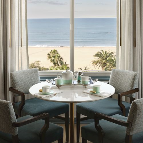 A round table set for tea with four chairs by a window, overlooking a beach and ocean view with palm trees in the distance.