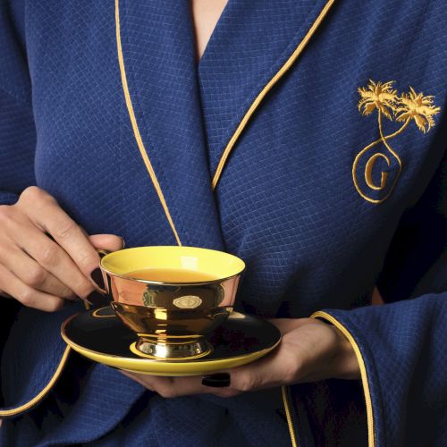 A person in a blue robe with gold trim is holding a golden teacup and saucer, featuring a palm tree design and a letter "G" monogram.
