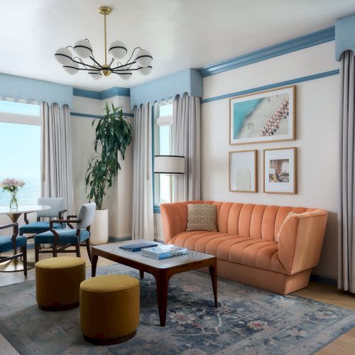 Bright seaside living room with orange sofa, coffee table, blue curtains, wall art, chandelier, and ocean-view windows.