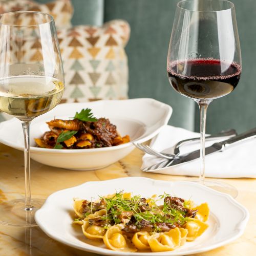 Two plates of pasta dishes served with a glass of white wine and a glass of red wine on a table, with utensils and napkin.