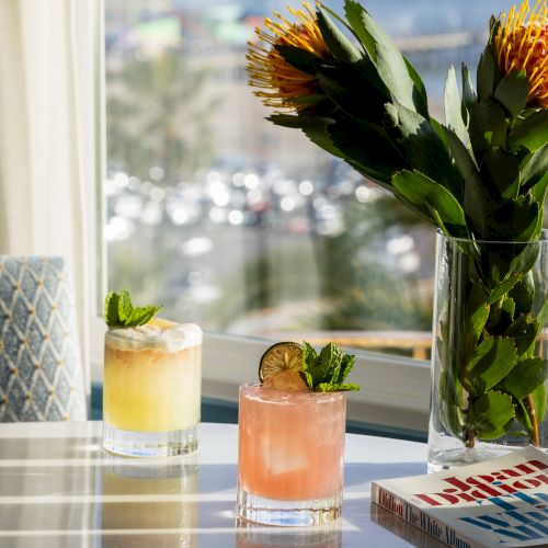 Two cocktails with garnishes sit on a table near a vase of flowers and a book, with a window view in the background.