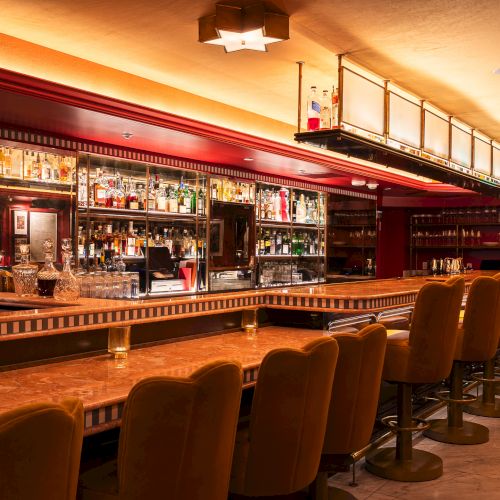 A stylish, warmly-lit bar with plush seating, polished counters, and shelves stocked with a variety of spirits and glasses.