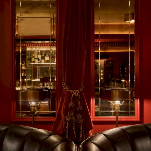 A dimly lit room features leather seating, red curtains, and mirrors reflecting a well-stocked bar in the background.