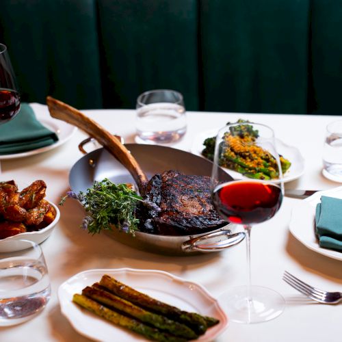 A table setting with steak, vegetables, and wine. Plates, glasses, and utensils are arranged on a white tablecloth.