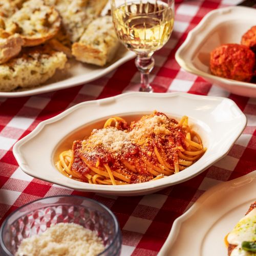 The image shows a spread of Italian dishes, including pasta, meatballs, salad, garlic bread, and wine on a red checkered tablecloth.