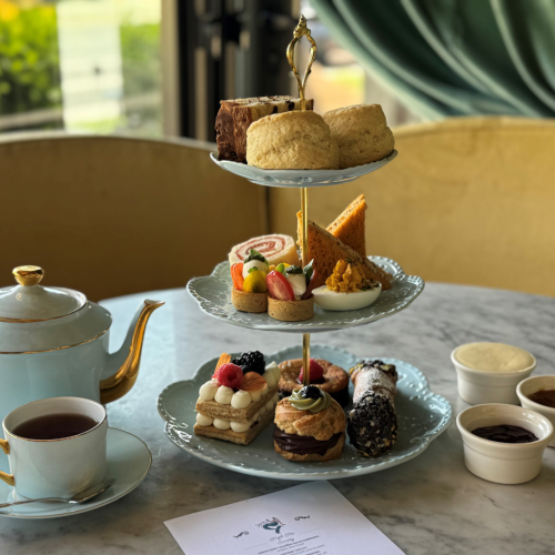 The image shows a three-tiered tray with assorted pastries and scones, alongside a teapot, teacup, and small condiment bowls on a marble table.