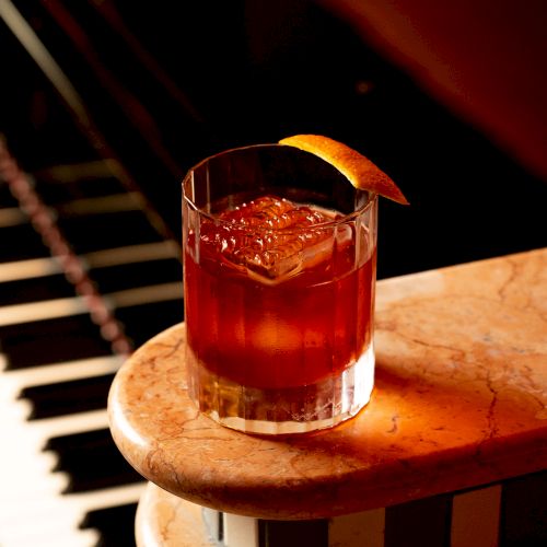 A cocktail with an orange peel garnish sits on a marble surface, next to a piano keyboard.