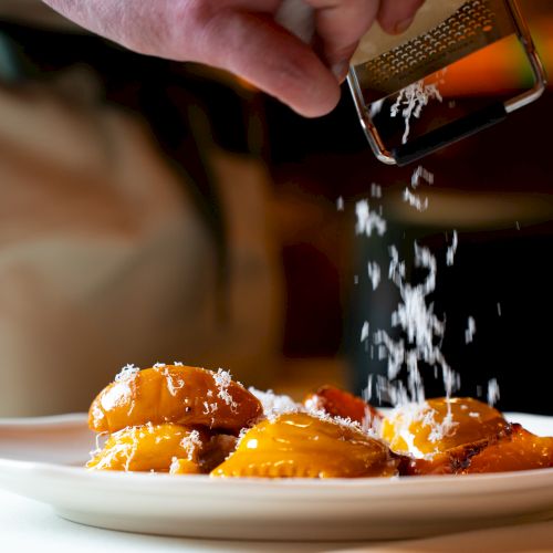 A hand grates cheese over a plate of ravioli, adding a finishing touch to the dish.