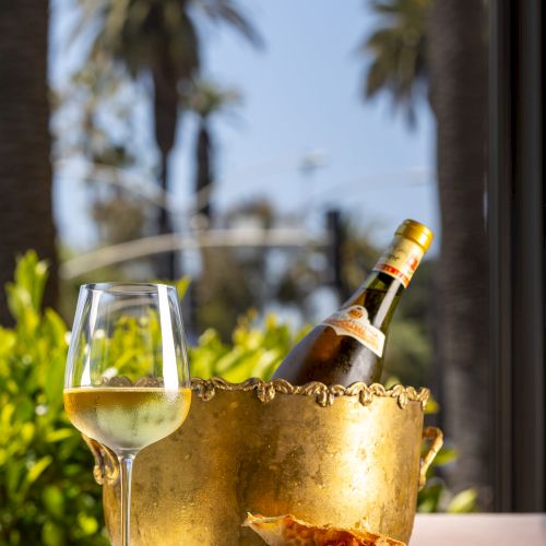 A chilled wine bottle in a bucket, a glass of white wine, and a plate with food are set on a table outdoors against a backdrop of palm trees.