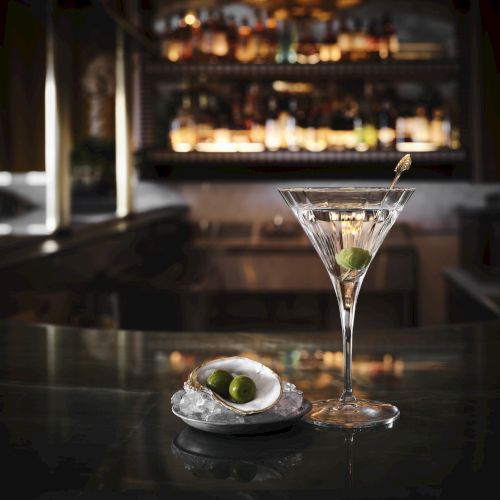 A martini with an olive skewered on a stick is on a bar counter, next to an oyster shell containing an olive. A dimly lit bar is in the background.