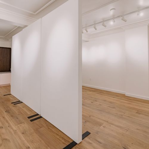 The image shows an empty art gallery room with wooden floors, white walls, and a colorful artwork in the distance on the left.