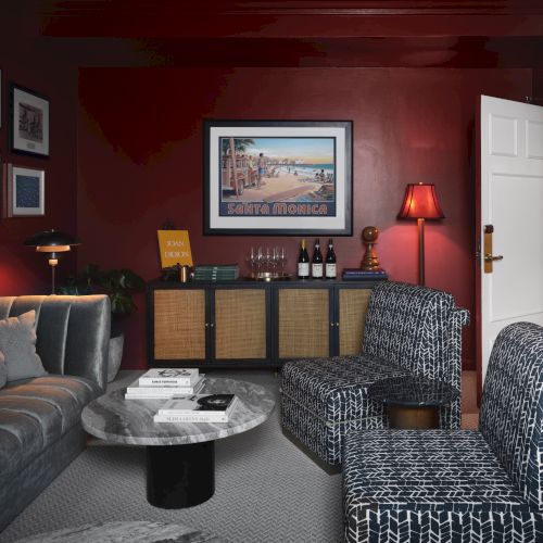 A cozy living room with red walls, gray sofa, patterned chairs, a round marble table, and wall art. It also features a cabinet and a table lamp.