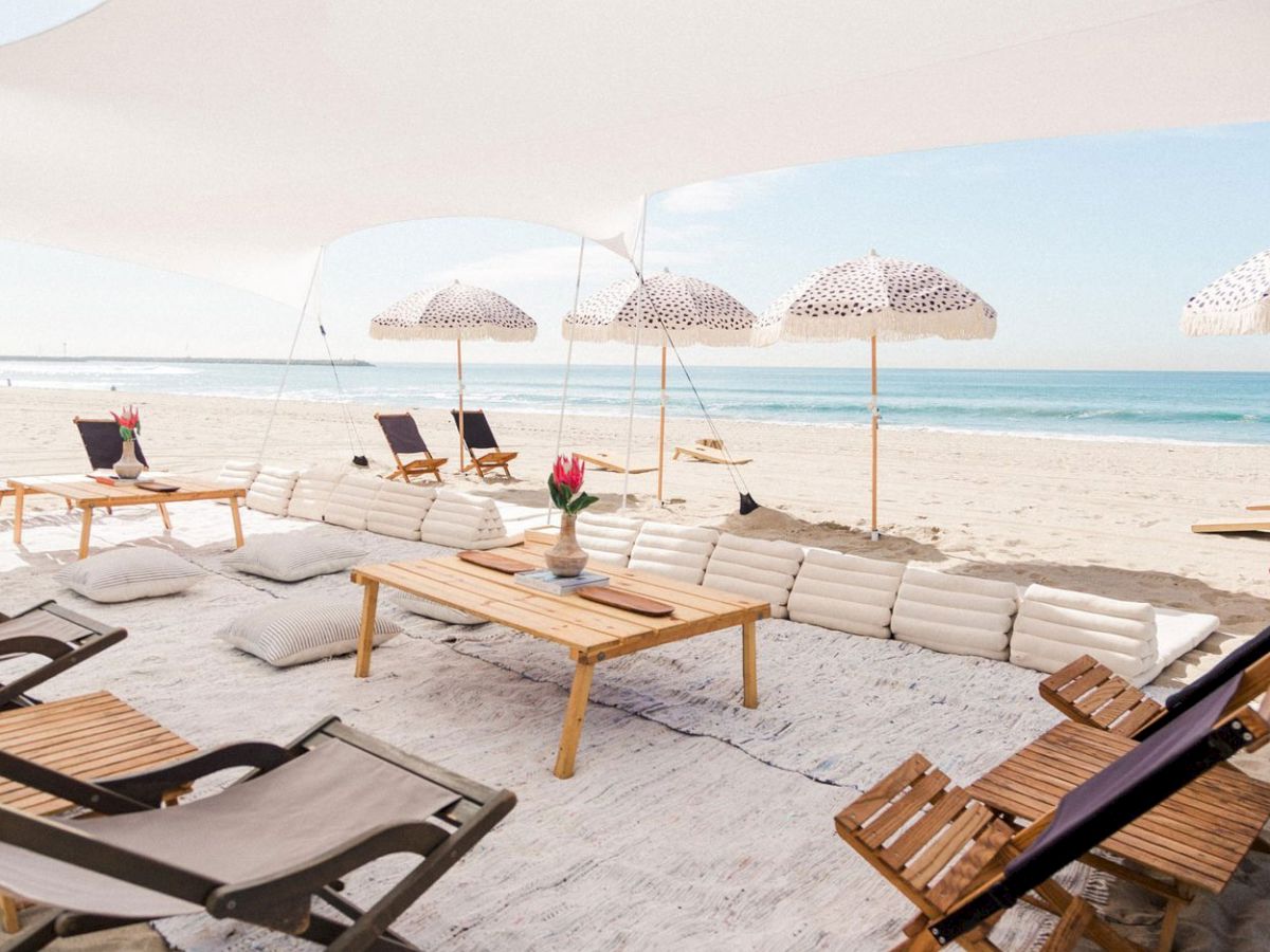 A beach setup with wooden tables, cushioned seating, and parasols under a canopy, offering a relaxing seaside view with deck chairs.