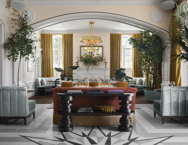 A luxurious hotel lobby with a grand central desk, emerald-green chairs, tall gold curtains, and plants framing a sunlit, elegant seating area.