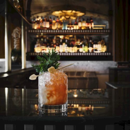 A cocktail on a dark bar counter with ice, citrus hues, a sprig of mint, and a blurred well-lit liquor shelves in the background.