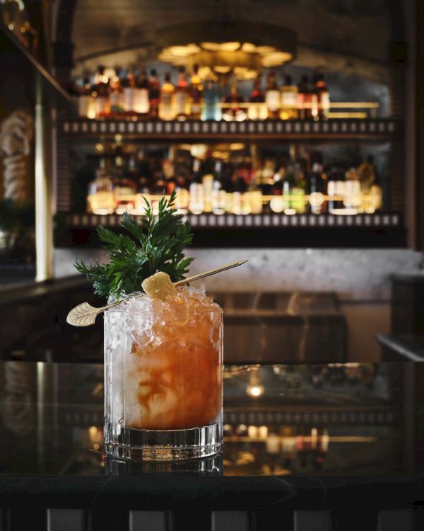 A cocktail on a dark bar counter with ice, citrus hues, a sprig of mint, and a blurred well-lit liquor shelves in the background.