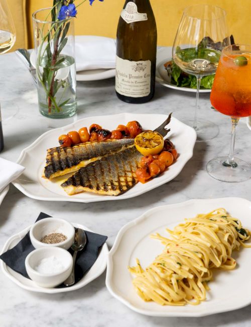 A table setting with grilled fish, pasta, a bottle of wine, a cocktail, and flowers in a vase on a marble surface.