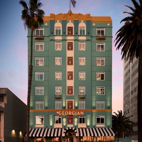 Georgian-style turquoise hotel with white trim, striped awnings, palm trees, and a flag on top; &ldquo;GEORGIAN&rdquo; signage; warm sunset glow. Ends with a period.