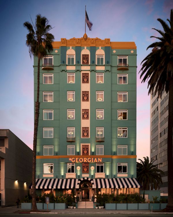 Georgian-style turquoise hotel with white trim, striped awnings, palm trees, and a flag on top; &ldquo;GEORGIAN&rdquo; signage; warm sunset glow. Ends with a period.