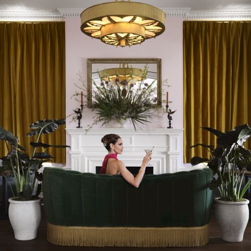 A woman in a red dress sits on a green sofa in a stylish living room with gold curtains, potted plants, and a floral centerpiece above a fireplace.