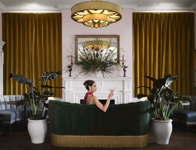 A woman in a red dress sits on a green sofa in a stylish living room with gold curtains, potted plants, and a floral centerpiece above a fireplace.