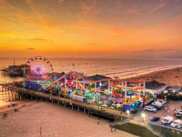 A brightly lit amusement park on a pier at sunset, overlooking a beach and ocean with people strolling, cars parked nearby.