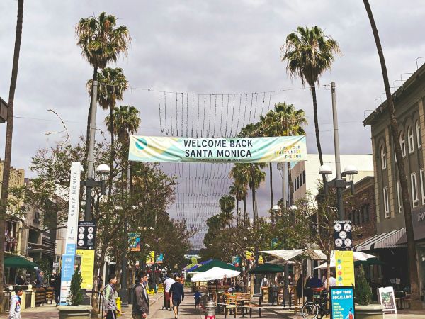 A pedestrian street lined with palm trees, outdoor seating, and shops under a cloudy sky.