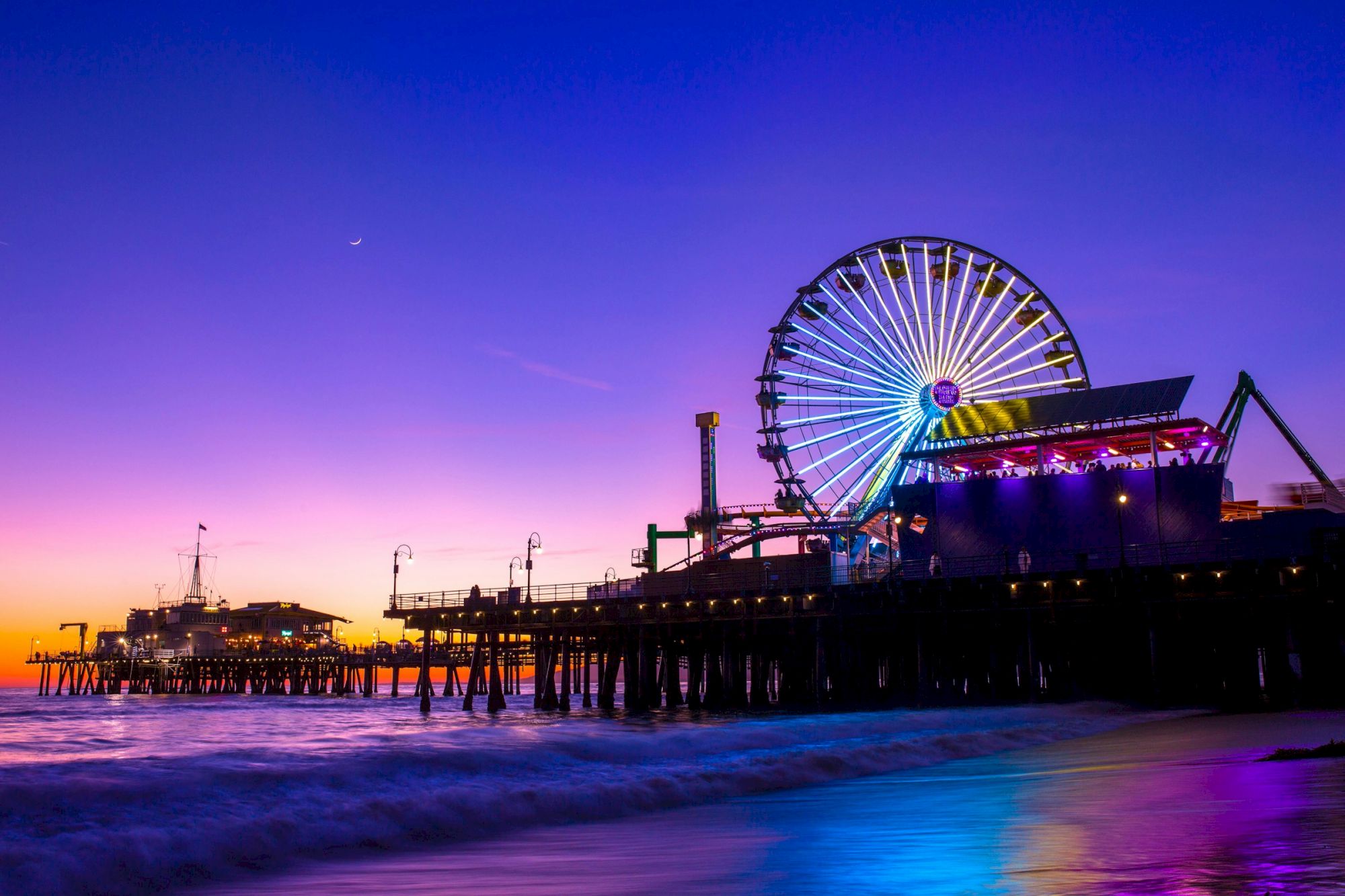 A vibrant sunset over a pier with a Ferris wheel lit up, reflecting colorful lights on the ocean waves.