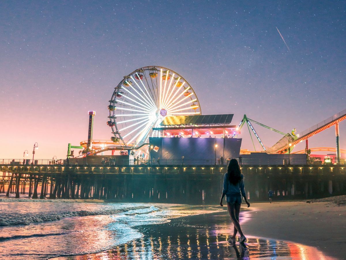 A person walks towards a lit-up Ferris wheel on a pier at twilight, with reflections shimmering on the wet sand.