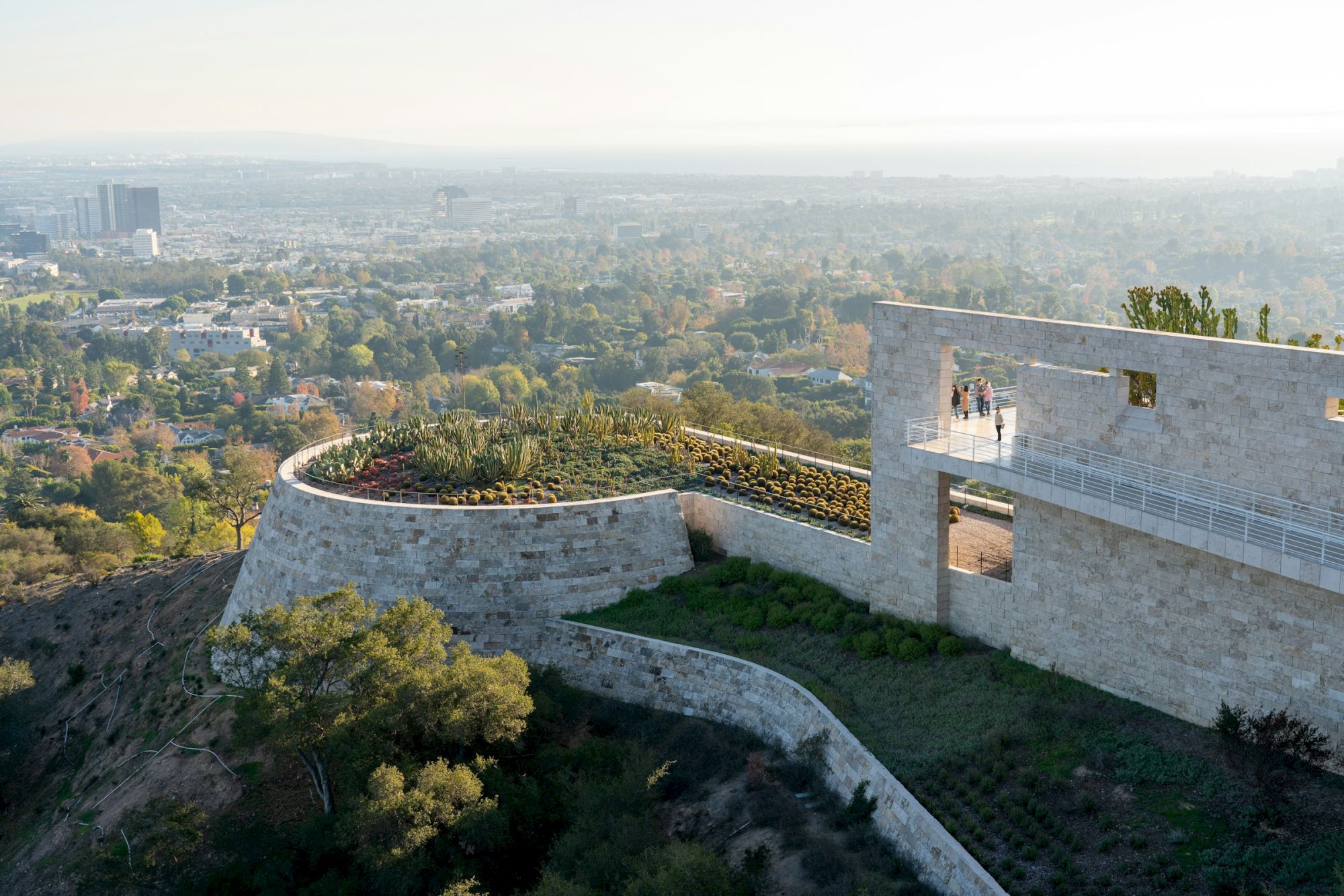 A modern architectural structure overlooks a sprawling cityscape with greenery and distant buildings under a clear sky.