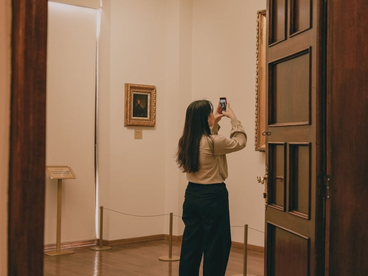 A person is taking a photo of an artwork in a gallery. The interior is warmly lit, and the walls display framed paintings.