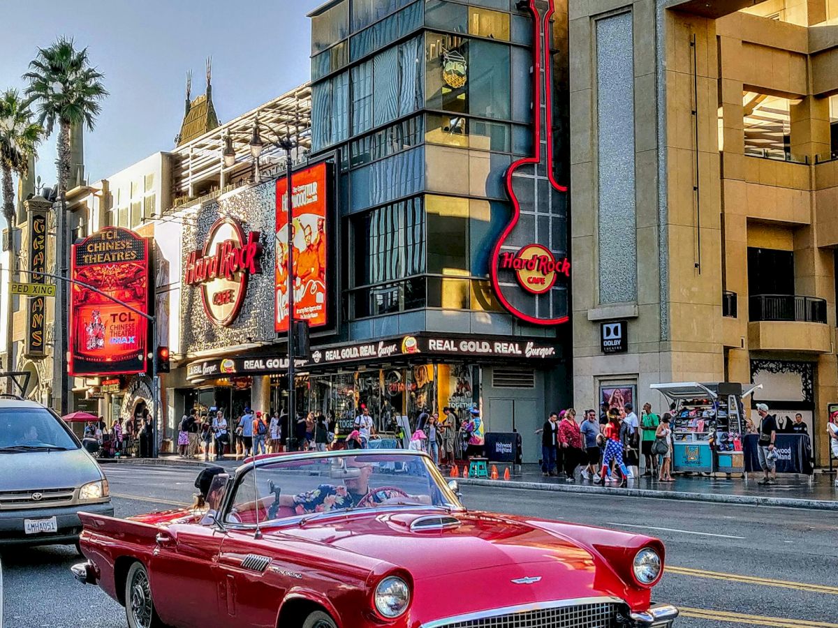 A red vintage convertible drives past lively buildings and a crowd on a city street, with iconic signs and palm trees in the background.