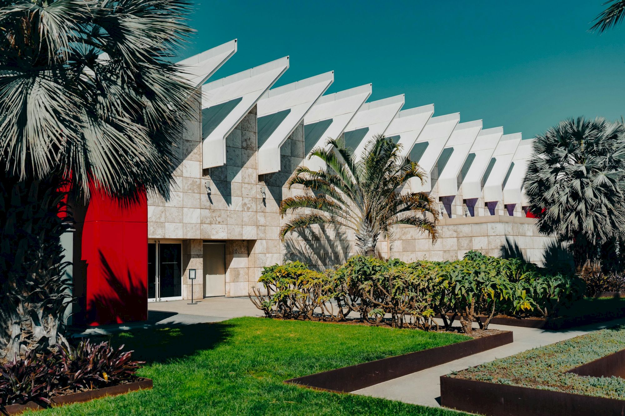 A modern building with a geometric roof, surrounded by palm trees and greenery, under a clear blue sky.