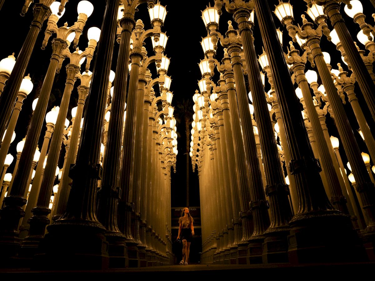 A person stands among rows of illuminated streetlamps at night, creating a symmetrical and artistic visual effect.