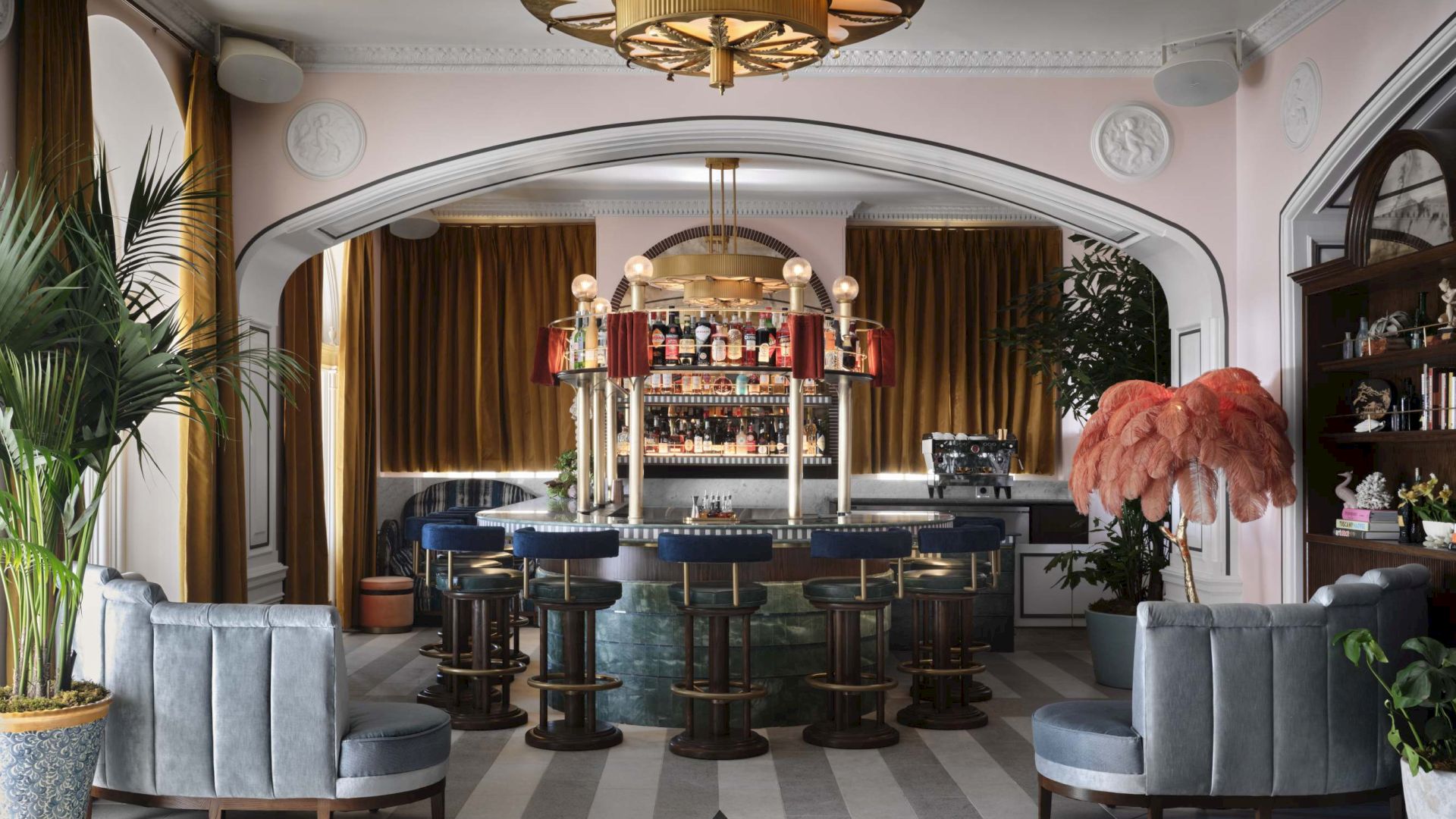 A stylish bar interior with an ornate ceiling light, patterned floor, plush seating, and decorative plants and feathers.