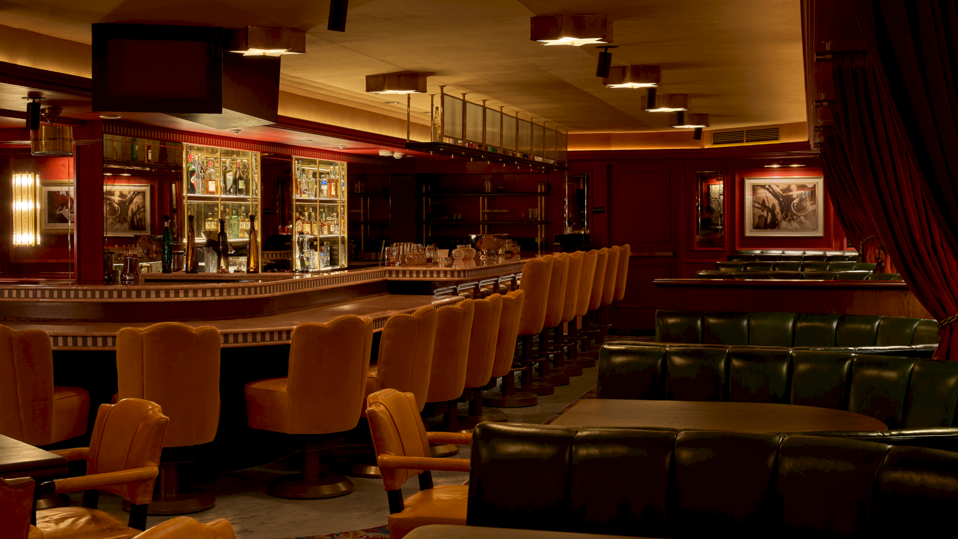 A cozy hotel bar lounge with a back-lit liquor shelf, cushioned seats, framed photos, and warm ceiling lights.