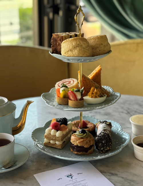 Afternoon tea setup with a tiered stand of pastries, sandwiches, scones; teapot and cup on the side.