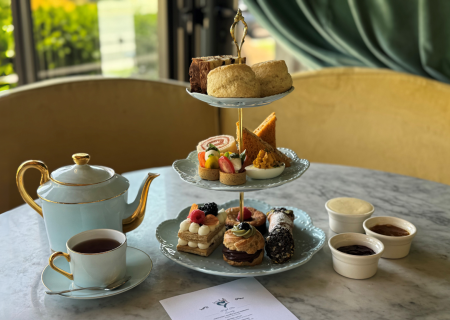 A tea setup with a teapot, cups, and a pastry on a plate beside a small tiered stand, elegant afternoon tea scene.