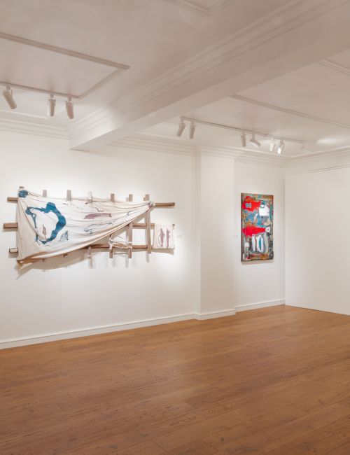 An art gallery with white walls and wooden floors, featuring several abstract mixed-media paintings and sculptural pieces.