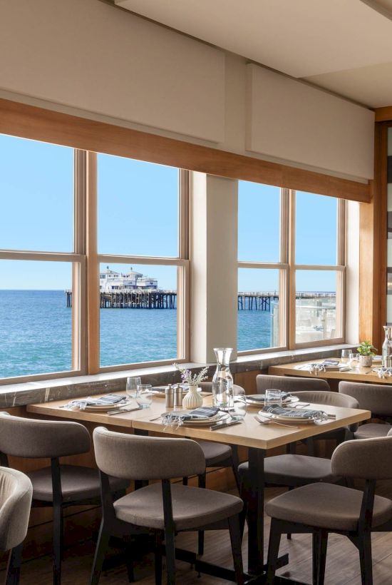 A bright coastal restaurant with large windows, wooden tables, and cushioned chairs facing the sea. The calm ocean and pier are visible outside.