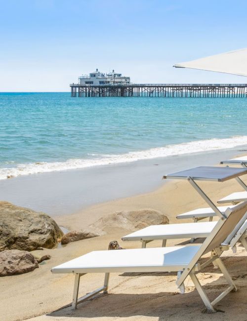 Rows of white lounge chairs under umbrellas on a sunny sandy beach, with a wooden pier stretching into calm blue water.